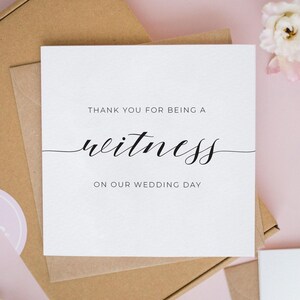 以下が含まれることがあります： THANK YOU FOR BEING A witness ON OUR WEDDING DAYという黒い文字が印刷された正方形の白いグリーティングカード。witnessという単語は装飾的な筆記体で書かれており、他の単語はシンプルなサンセリフフォントで構成されています。