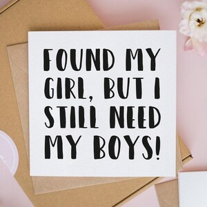 May include: A square white greeting card with black text that reads, "FOUND MY GIRL, BUT I STILL NEED MY BOYS!" The card is positioned on top of a brown cardboard envelope, with a small white flower visible in the upper right corner of the frame.