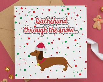 May include: A square greeting card with a white background featuring red and green confetti dots. The text reads Dachshund through the snow in red letters. A brown dachshund wearing a red and white Santa hat and a red collar is centered.