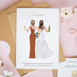 May include: A bridesmaid proposal card with an illustration of two women, one in a rust-coloured dress and the other in a white wedding gown. The card is personalised with the name "Lauren" and the text "Act surprised like you had no idea this was happening. Will you be my bridesmaid?"