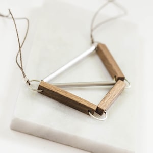 Puede incluir: Un collar geométrico hecho de madera y plata. El collar presenta una forma triangular con una barra de plata que cruza el centro. El collar está suspendido de un cordón marrón.