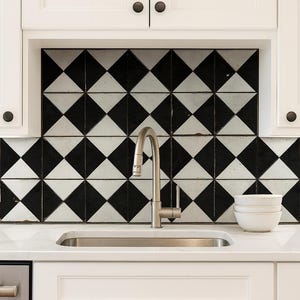 May include: Kitchen with a black and white diamond tile backsplash. Stainless steel tap over a stainless steel sink. White cabinets with black knobs. Two white bowls on the countertop.