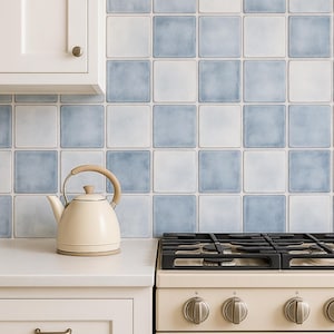 Puede incluir: Una tetera color crema con un asa de madera se encuentra sobre una encimera blanca junto a una estufa de gas. El protector contra salpicaduras presenta baldosas cuadradas en tonos azules y blancos alternos, creando una estética de cocina clásica.