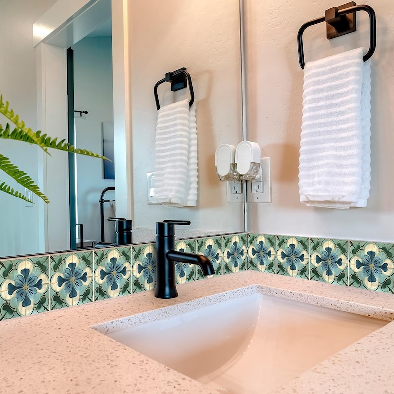 Puede incluir: Un lavabo de ba&ntilde;o blanco con un grifo negro y una toalla blanca colgada de un toallero negro. El lavabo est&aacute; colocado sobre un fondo de azulejos con motivos verdes y azules.