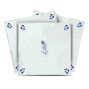 Peut inclure: Plusieurs carreaux de céramique carrés blanc cassé, d'aspect vieilli. Chaque carreau présente un motif floral bleu dans les coins et une illustration botanique centrale. Les carreaux sont disposés pour mettre en valeur le design.