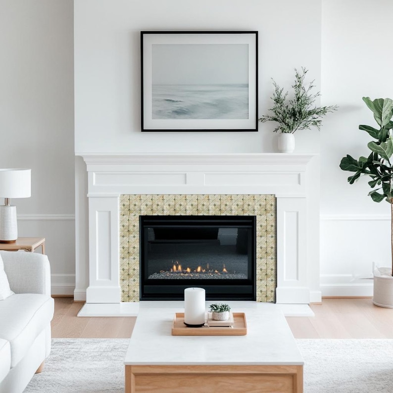 May include: A white fireplace with a black framed picture above it. The fireplace has a decorative tile surround and a lit fire. A white coffee table with a wooden base sits in front of the fireplace, with a candle and tray on top.