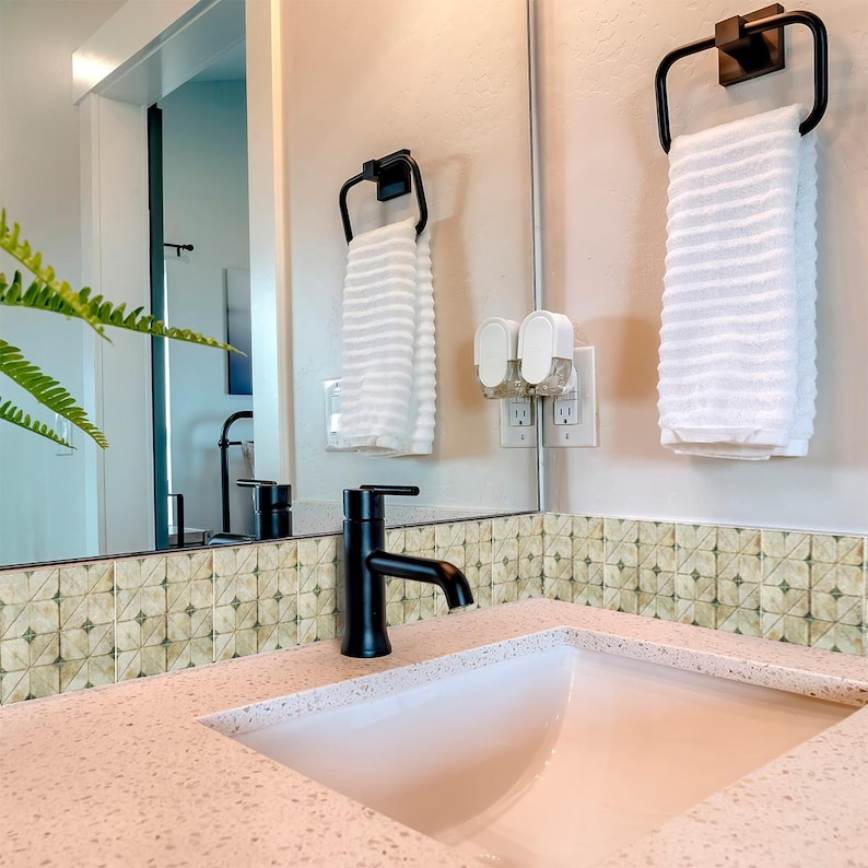 May include: A modern bathroom sink with a black faucet and a white countertop. Two black towel rings hold white towels. The mirror reflects the sink and a glimpse of a fern. The wall has a decorative tile border.