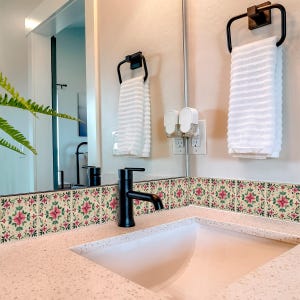 May include: A bathroom sink with a white countertop and a black faucet. The backsplash is decorated with pink and green floral tile stickers. A white towel hangs on a black towel rack.
