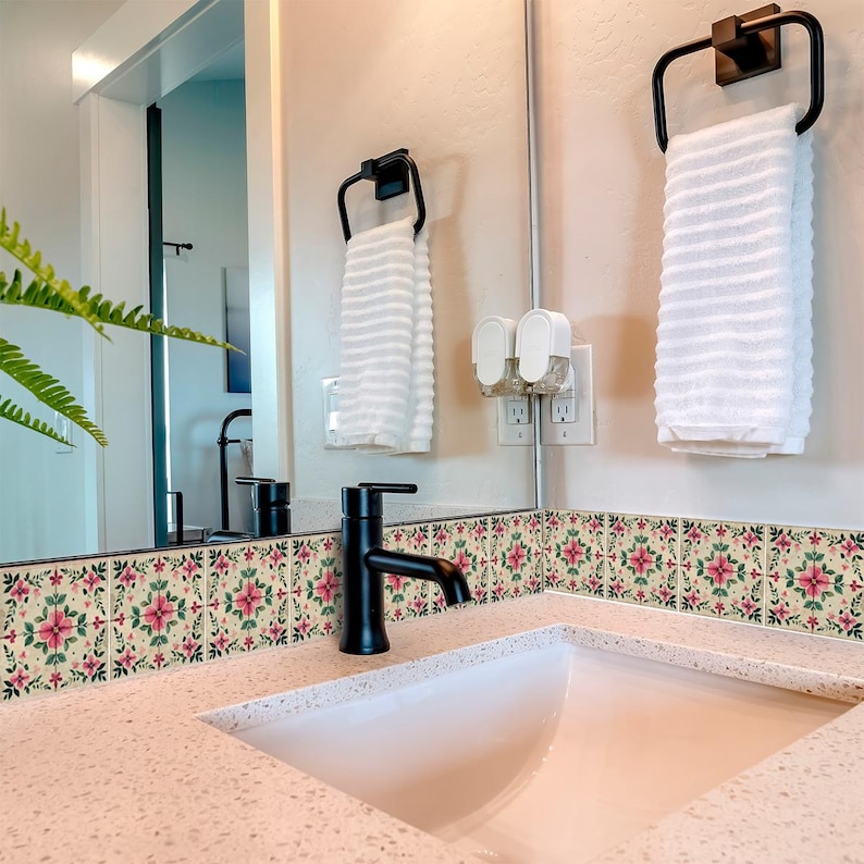May include: A bathroom sink with a white countertop and a black faucet. The backsplash is decorated with pink and green floral tile stickers. A white towel hangs on a black towel rack.