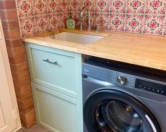 May include: A laundry room setup featuring a light green cabinet with a black handle, a light wood countertop, and a white sink with a chrome faucet. The wall is covered in pink and green floral patterned tiles above a red brick wall section.