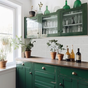 Puede incluir: Una escena de cocina con gabinetes verde oscuro y una encimera de madera. Botellas de vidrio y plantas en macetas están dispuestas en la encimera y los estantes. Los gabinetes tienen puertas de vidrio y una ventana a la izquierda.