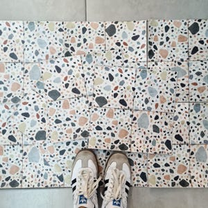 Puede incluir: Primer plano de un suelo de baldosas con fondo blanco y pequeñas piedras de colores incrustadas en la superficie. Dos zapatillas blancas con rayas negras colocadas en el suelo.