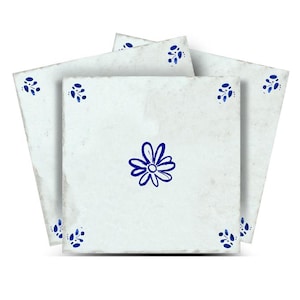 Peut inclure: Plusieurs carreaux de céramique carrés, blanc cassé, d'aspect vieilli. Chaque carreau présente une fleur bleue dessinée à la main au centre et de petits motifs floraux bleus dans les coins. Les carreaux sont disposés pour mettre en valeur le motif.