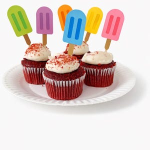 May include: A white plate holds six red velvet cupcakes topped with white frosting and popsicle-shaped toppers in various colors. The cupcakes are arranged on a white plate, creating a festive and colorful dessert display.