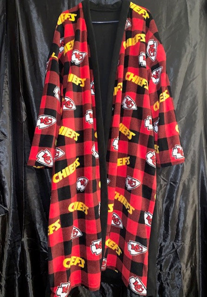 May include: A red and black fleece robe with a chequered pattern and the word "Chiefs" repeated throughout. The robe has a black inner lining and a shawl collar.