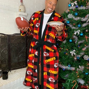 May include: A person wearing a red and black plaid robe with the word "Chiefs" printed in yellow. The robe has a black sash and is decorated with the Kansas City Chiefs logo. The person is holding a football and a plate with a pancake on it.