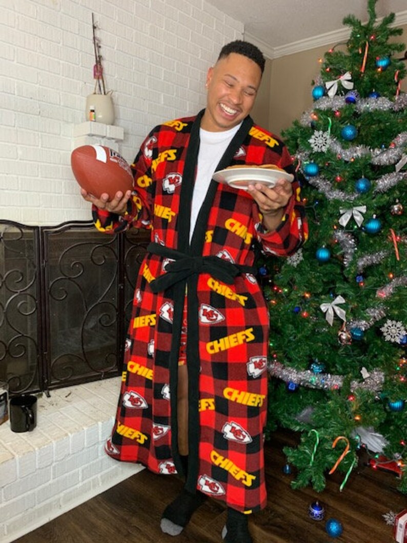 May include: A person wearing a red and black plaid robe with the word "Chiefs" printed in yellow. The robe has a black sash and is decorated with the Kansas City Chiefs logo. The person is holding a football and a plate with a pancake on it.