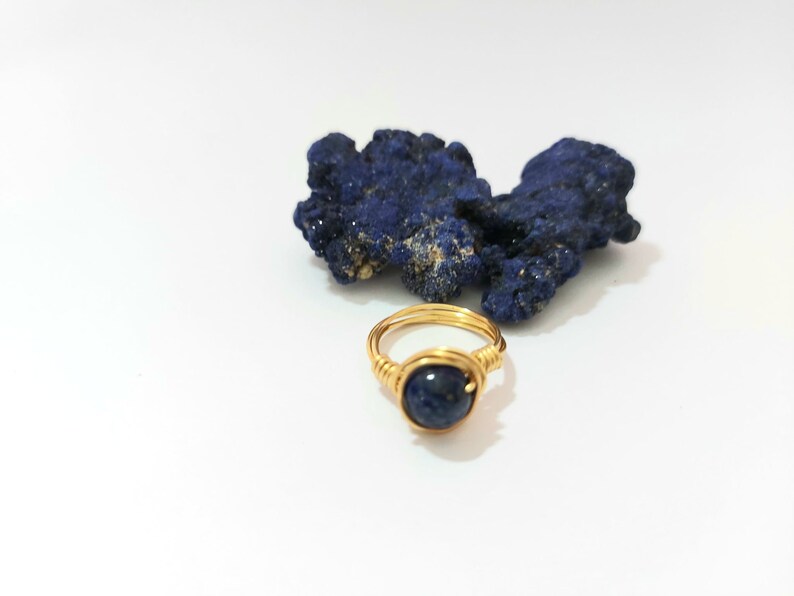 K&ouml;nnte beinhalten: Ein goldfarbener Ring, der mit Draht umwickelt ist und einen dunkelblauen Edelstein tr&auml;gt. Der Ring liegt auf einer wei&szlig;en Oberfl&auml;che neben einem St&uuml;ck blauen Azurite.
