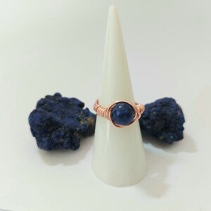 K&ouml;nnte beinhalten: Ein Ring aus Kupferdraht mit einem dunkelblauen Edelstein. Der Ring wird auf einem wei&szlig;en Kegel pr&auml;sentiert.