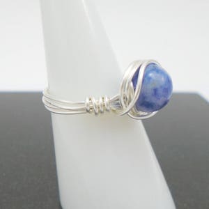 May include: A silver wire wrapped ring with a blue gemstone. The ring is on a white display stand.