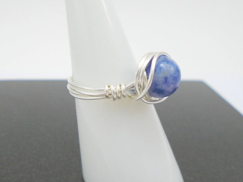 May include: A silver wire wrapped ring with a blue gemstone. The ring is on a white display stand.