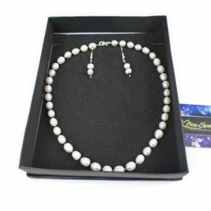 K&ouml;nnte beinhalten: Ein Perlenschmuck-Set in einer schwarzen Schachtel. Die Halskette besteht aus grauen Perlen und die Ohrringe sind ein passendes Paar grauer Perlenstecker.