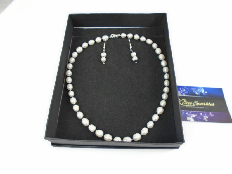 K&ouml;nnte beinhalten: Ein Perlenschmuck-Set in einer schwarzen Schachtel. Die Halskette besteht aus grauen Perlen und die Ohrringe sind ein passendes Paar grauer Perlenstecker.