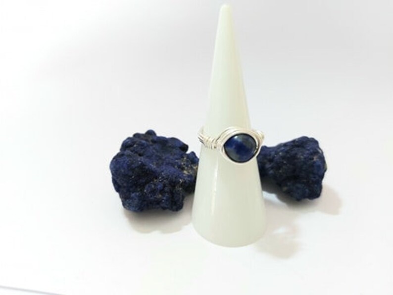 K&ouml;nnte beinhalten: Ein Ring aus Silberdraht mit einem blauen Edelstein. Der Ring wird auf einem wei&szlig;en Kegel pr&auml;sentiert.
