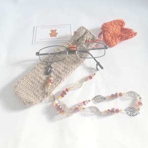 May include: A beige knitted glasses case with a brown and black pair of glasses inside. The glasses have a beaded chain with silver leaf charms and red and orange beads.