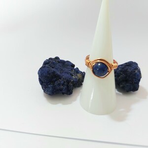 K&ouml;nnte beinhalten: Ein Ring aus Kupferdraht, der mit einem dunkelblauen Edelstein umwickelt ist. Der Ring wird auf einem wei&szlig;en Kegel mit zwei St&uuml;cken blauen Azurits pr&auml;sentiert.