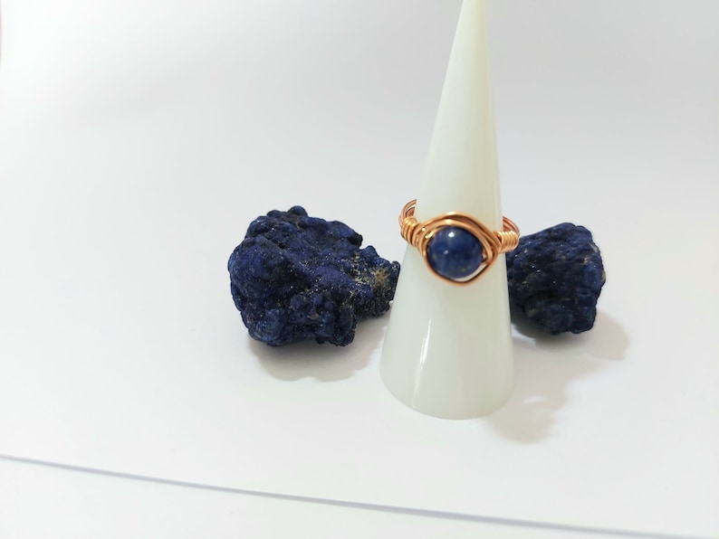 K&ouml;nnte beinhalten: Ein Ring aus Kupferdraht, der mit einem dunkelblauen Edelstein umwickelt ist. Der Ring wird auf einem wei&szlig;en Kegel mit zwei St&uuml;cken blauen Azurits pr&auml;sentiert.