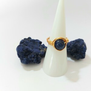 K&ouml;nnte beinhalten: Ein golddrahtumwickelter Ring mit einem dunkelblauen Edelstein. Der Ring wird auf einem wei&szlig;en Kegel pr&auml;sentiert.