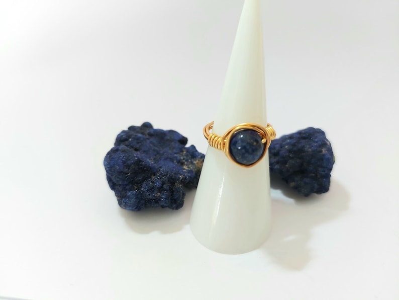 K&ouml;nnte beinhalten: Ein golddrahtumwickelter Ring mit einem dunkelblauen Edelstein. Der Ring wird auf einem wei&szlig;en Kegel pr&auml;sentiert.