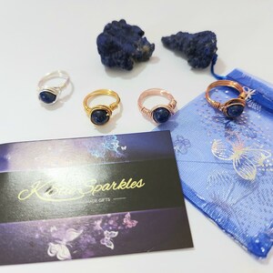 K&ouml;nnte beinhalten: Vier drahtgewickelte Ringe mit blauen Edelsteineins&auml;tzen. Die Ringe werden auf einer wei&szlig;en Oberfl&auml;che mit einer blauen und wei&szlig;en gemusterten Stofftasche und einem kleinen St&uuml;ck blauen Gesteins pr&auml;sentiert.