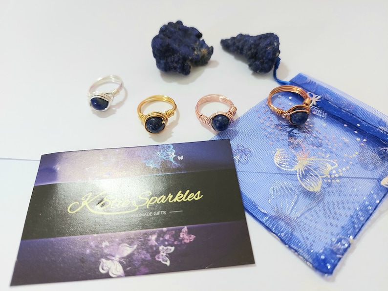 K&ouml;nnte beinhalten: Vier drahtgewickelte Ringe mit blauen Edelsteineins&auml;tzen. Die Ringe werden auf einer wei&szlig;en Oberfl&auml;che mit einer blauen und wei&szlig;en gemusterten Stofftasche und einem kleinen St&uuml;ck blauen Gesteins pr&auml;sentiert.