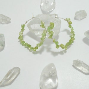 May include: A pair of silver hoop earrings with green gemstone accents. The earrings are made with small, irregular shaped green stones that are clustered together around the hoop.