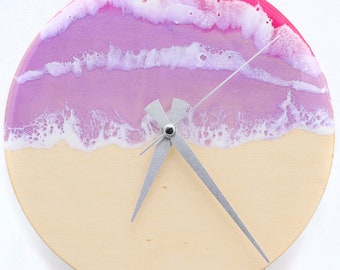 May include: A round wooden clock with a pink, purple, and white resin ocean wave design. The clock hands are silver.