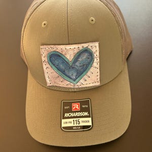 May include: Olive green trucker hat with a mesh back and a curved brim. The hat features a patch with a blue heart design. The Richardson 115 Trucker hat is MD/LG.