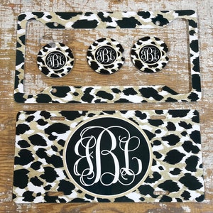 May include: A set of four black and white leopard print car accessories. The set includes a license plate frame, a key chain, and two cup coasters. The license plate frame and coasters feature a black circle with a white monogram in the center. The key chain is a round black and white leopard print disc with a white monogram in the center.