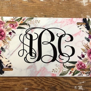 May include: A white personalized license plate with a pink and gray marble background. The plate features a floral design with pink, white, and purple flowers and a black monogram "JBC".