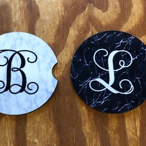 May include: Two round coasters with a marble design. One coaster is white with black marble veins and a black letter "B" with a swirl design. The other coaster is black with white marble veins and a white letter "L" with a swirl design.