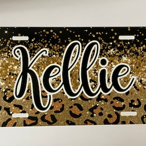 May include: A black and gold glitter license plate with a leopard print design and the name "Kellie" in black cursive lettering.