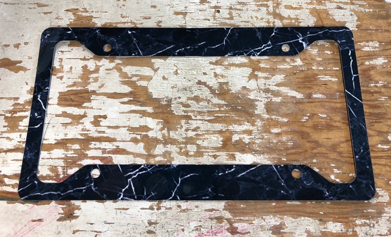 May include: Black and white marble license plate frame. The frame is made of plastic and has a smooth finish.