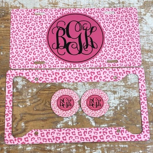 May include: Pink and black leopard print license plate frame with matching coasters. The frame and coasters feature a monogram in a circle.