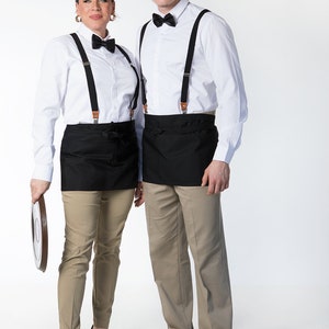May include: Two people wearing white shirts with black bow ties and black suspenders. They are both wearing black aprons and khaki pants. The woman is holding a brown tray.