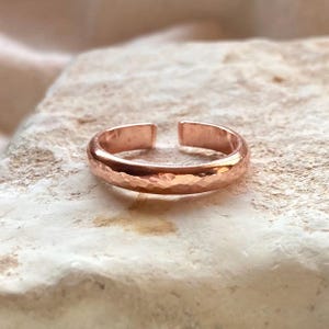 May include: A copper-coloured metal ring with an open band design. The surface of the ring features a distinct hammered texture, creating a series of small, reflective indentations across the outer circumference of the circular band.