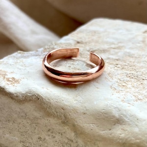 May include: A copper-coloured open ring. The ring is a simple band with a gap, allowing adjustment. It sits on a light-coloured, textured surface. The ring is made of a shiny, metallic material.