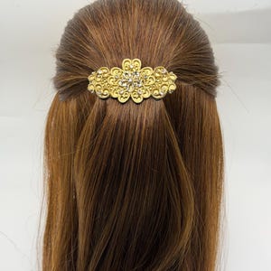 Könnte beinhalten: Eine goldfarbene Haarspange mit einem verzierten floralen Design und eingebetteten Strasssteinen. Die Spange ist an braunem Haar befestigt, das zu einer halben Hochsteckfrisur gestylt ist. Das Accessoire ist ein dekoratives Haarteil.