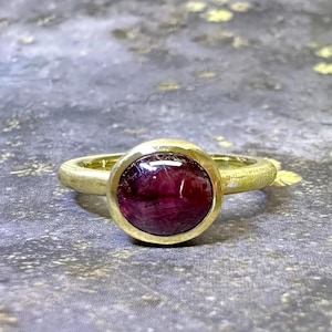 Könnte beinhalten: Ein goldener Ring mit einem großen, runden, tiefroten Edelstein. Der Ring hat ein schlichtes, elegantes Design mit einem glatten, polierten Band und einer Lünettenfassung, die den Edelstein sicher hält. Der Edelstein ist der Blickfang.