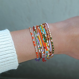 Peut inclure: Plusieurs bracelets de perles colorées empilés sur un poignet. Les bracelets présentent une variété de couleurs, notamment le rouge, l'orange, le vert et le violet. La personne porte également deux bagues en or. L'arrière-plan est de couleur neutre.